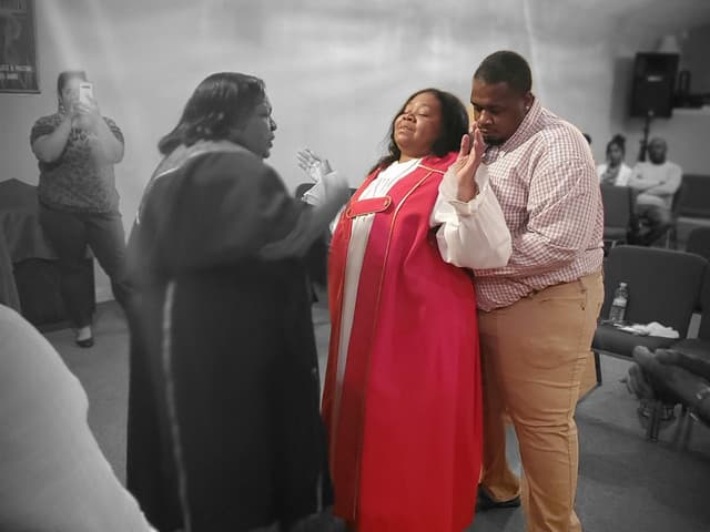 A commissioning prayer at Christ Family Church: two church members pray over a man in the sanctuary while another member records on her phone.