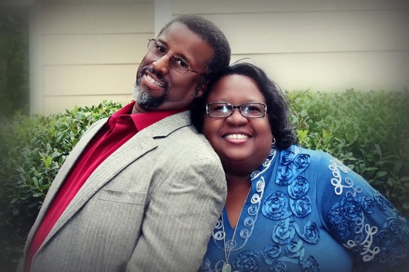Pastor Dennis Hood and Apostle Allice Hood standing together outdoors in front of a hedge, smiling, cheek to cheek.
