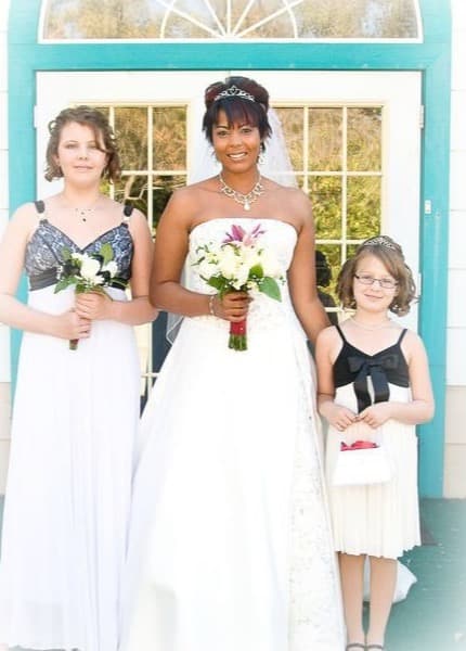 A bride holding a bouquet of lilies and roses, with two young bridesmaids, in front of arched white doors.